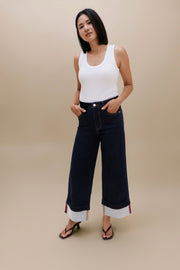 A woman modeling dark wash wide-leg cropped jeans with rolled cuffs, paired with a white tank top and black sandals, against a solid beige studio background.
