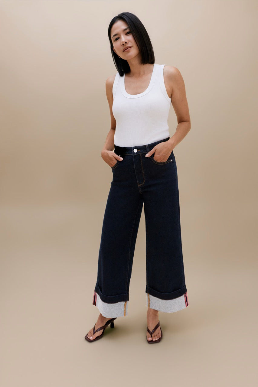 A woman modeling dark wash wide-leg cropped jeans with rolled cuffs, paired with a white tank top and black sandals, against a solid beige studio background.