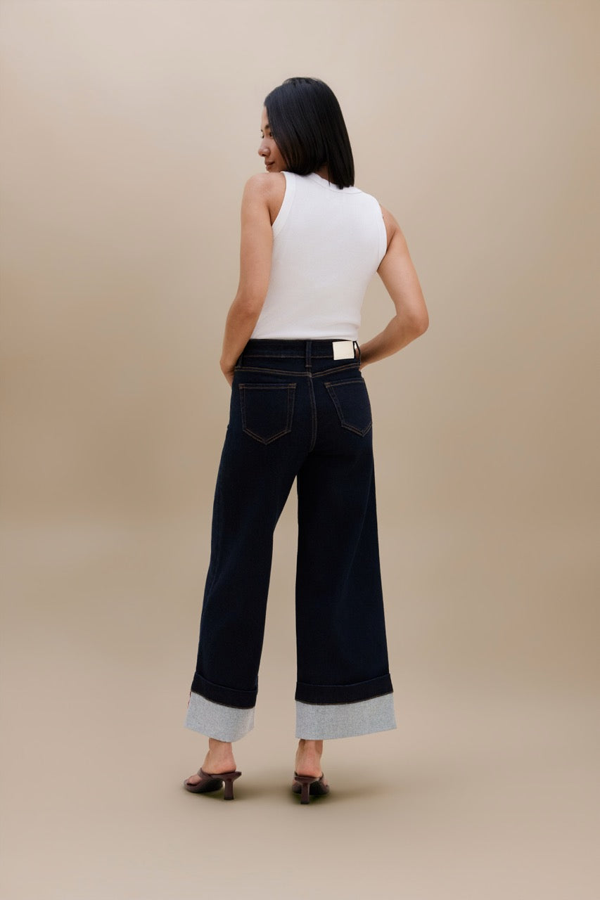 A woman is shown from behind wearing dark wide-leg cuffed jeans, a white sleeveless top, and black heels against a solid beige studio background.