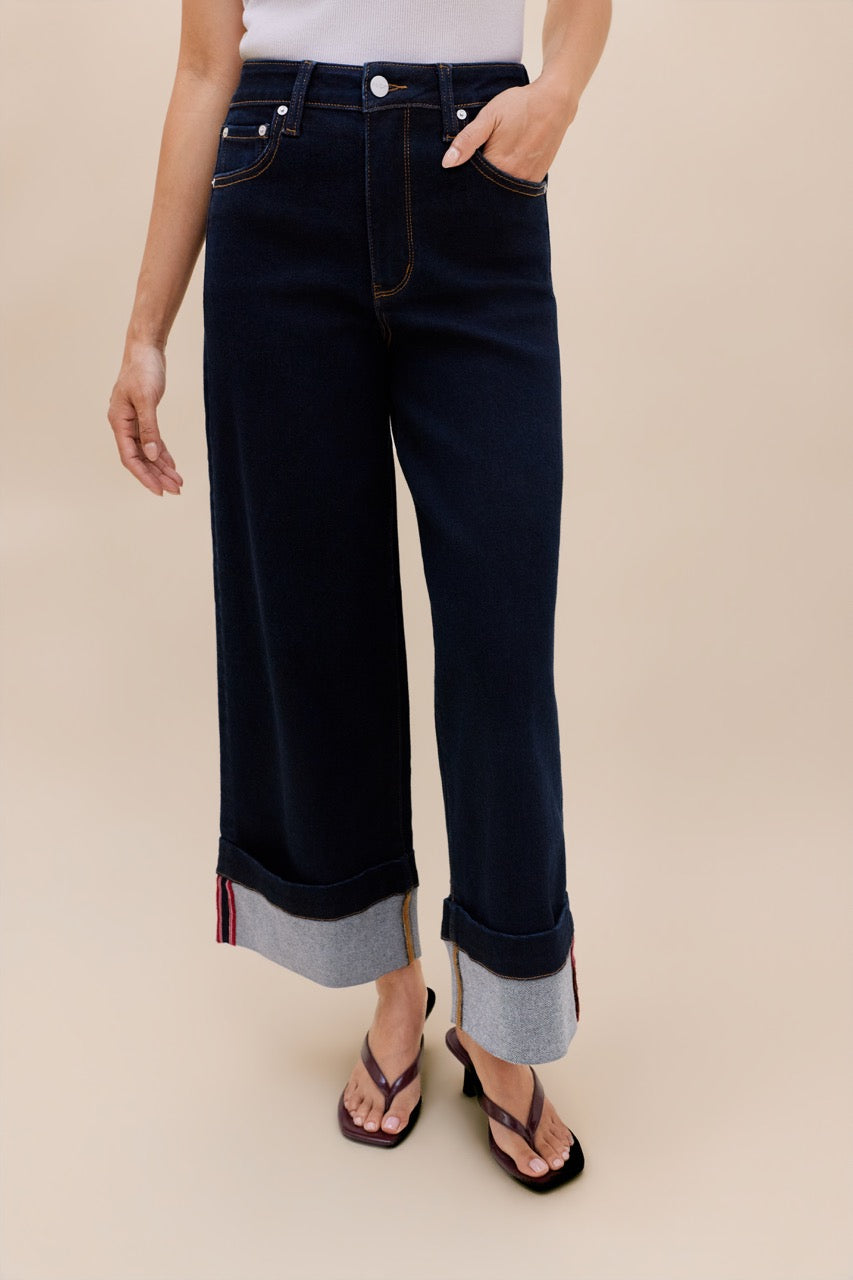 A model wearing dark navy cropped jeans with rolled cuffs showing a light gray inner lining, paired with black flip-flop sandals against a solid beige background.