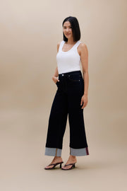 A woman modeling a pair of dark wide-leg cropped jeans with rolled cuffs showing a gray inner lining and thin red stripe detailing, paired with a white tank top and black sandals, against a solid beige studio background.