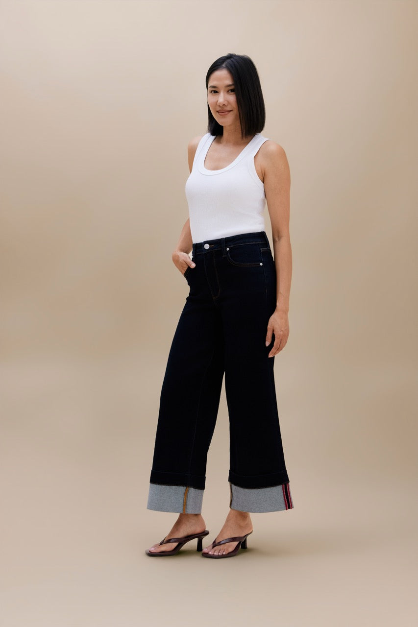 A woman modeling a pair of dark wide-leg cropped jeans with rolled cuffs showing a gray inner lining and thin red stripe detailing, paired with a white tank top and black sandals, against a solid beige studio background.