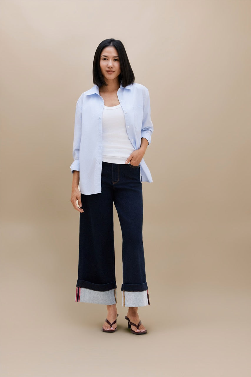 A woman stands against a plain beige studio background wearing a light blue button-up shirt over a white tank top, dark wide-leg cropped jeans with rolled cuffs, and dark sandals.