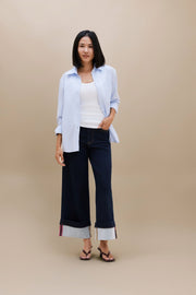 A woman stands against a plain beige studio background wearing a light blue button-up shirt over a white tank top, dark wide-leg cropped jeans with rolled cuffs, and dark sandals.
