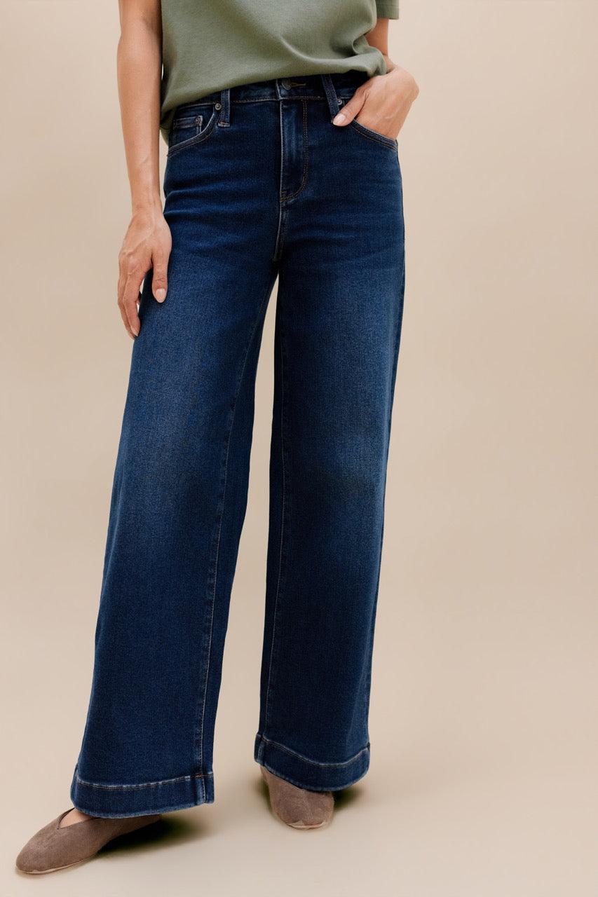 A person wearing dark blue wide-leg jeans with a light olive green top and taupe flats, standing against a solid beige studio background.