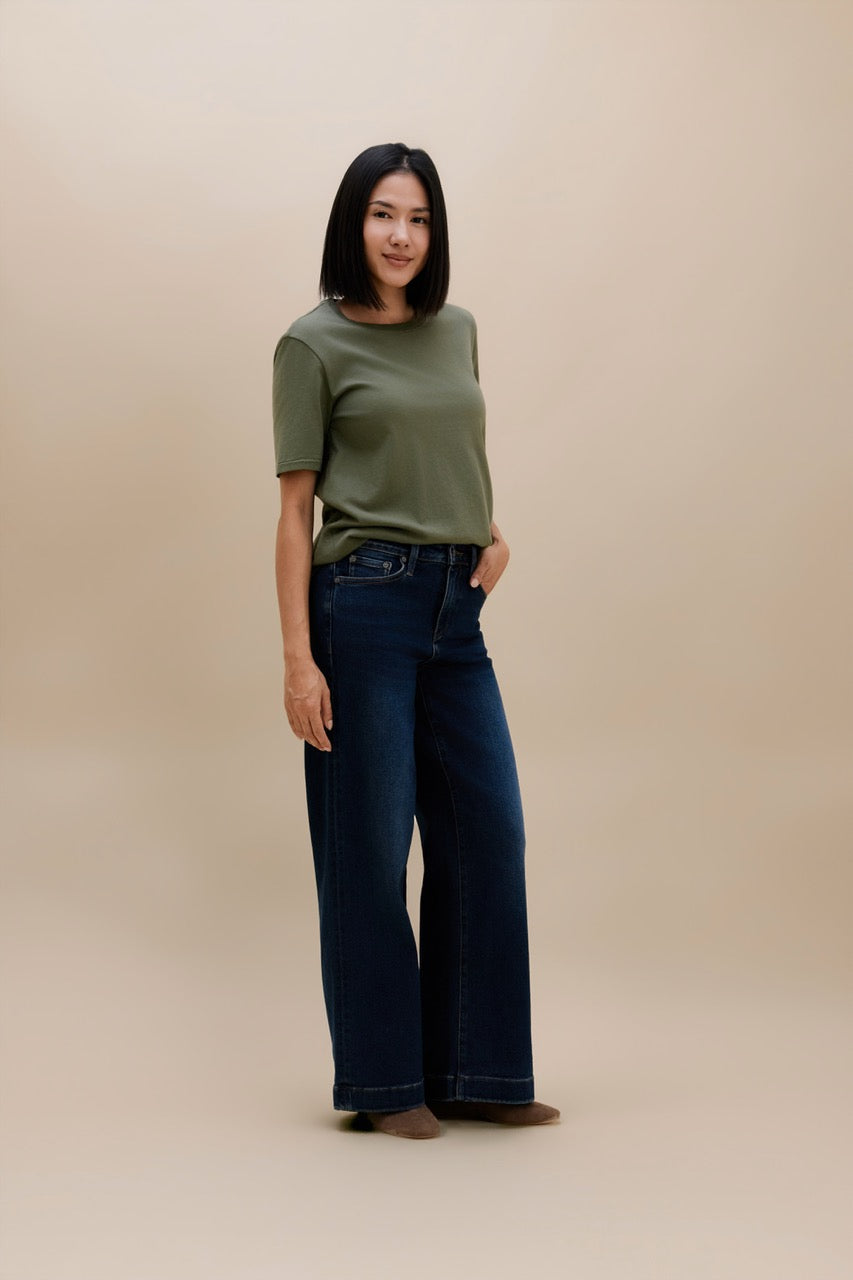 A woman modeling a short-sleeve olive green top and dark wide-leg blue jeans against a solid beige studio background.