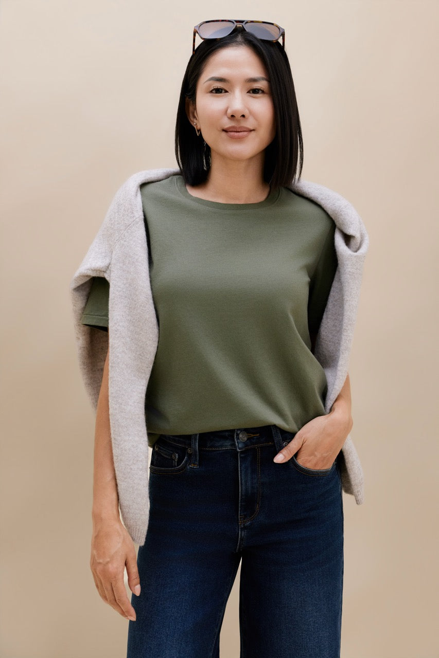A woman modeling an olive green crewneck t-shirt, styled with a light gray knit cardigan draped over her shoulders and dark blue jeans, against a solid beige background.