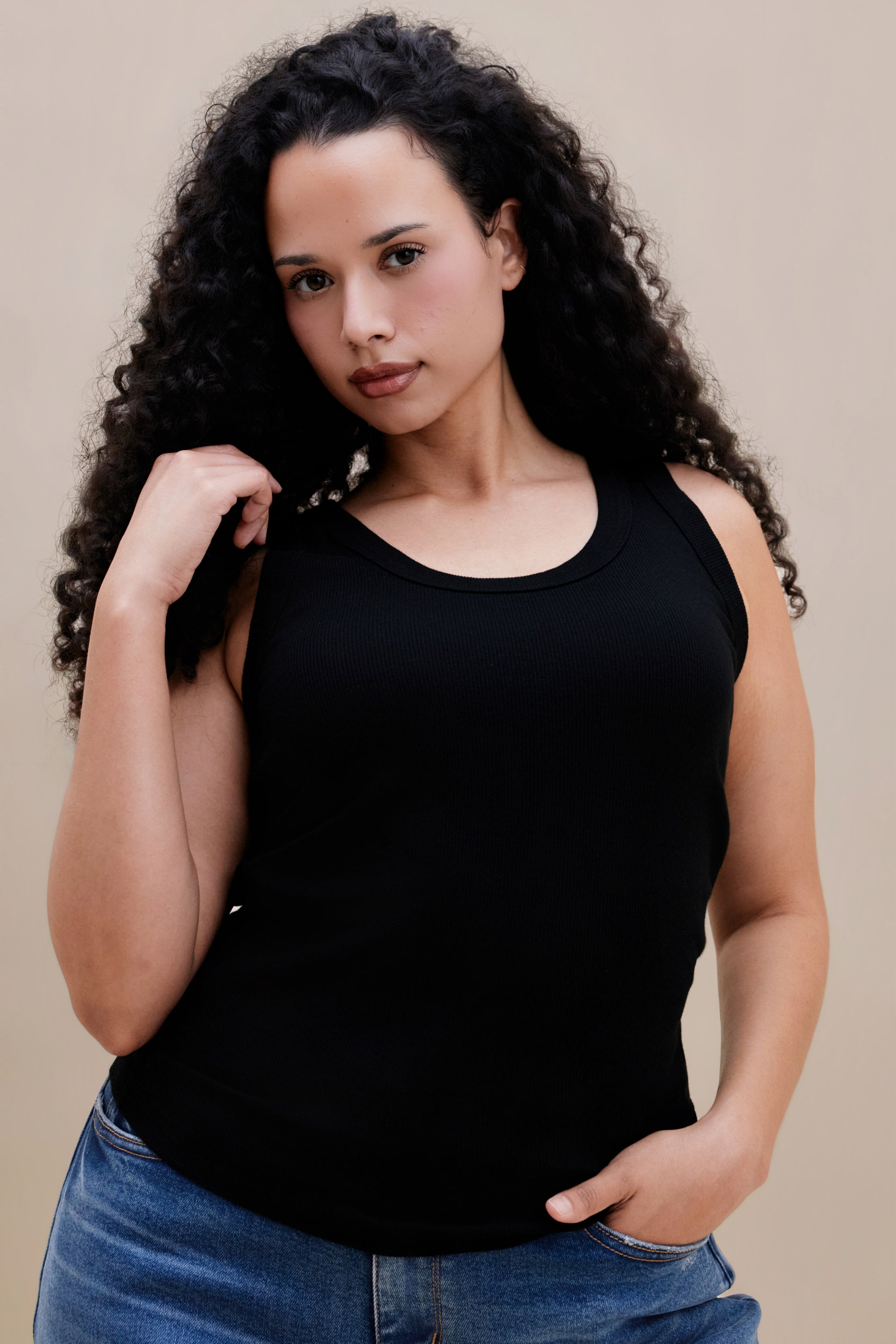 Woman wearing a black ribbed tank top and jeans against a beige background