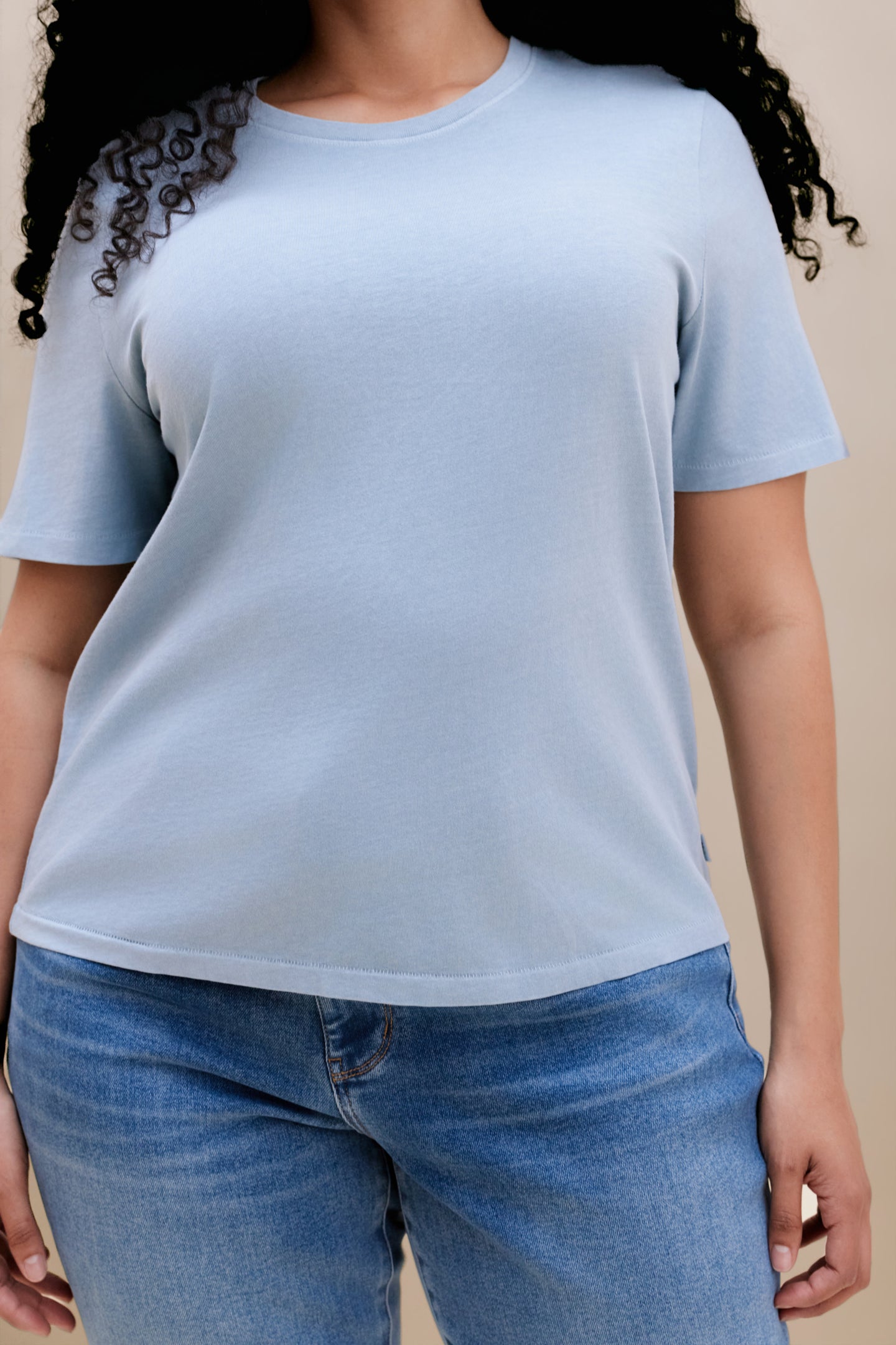 Close up of a woman wearing a blue crew neck tee and jeans with a beige background 
