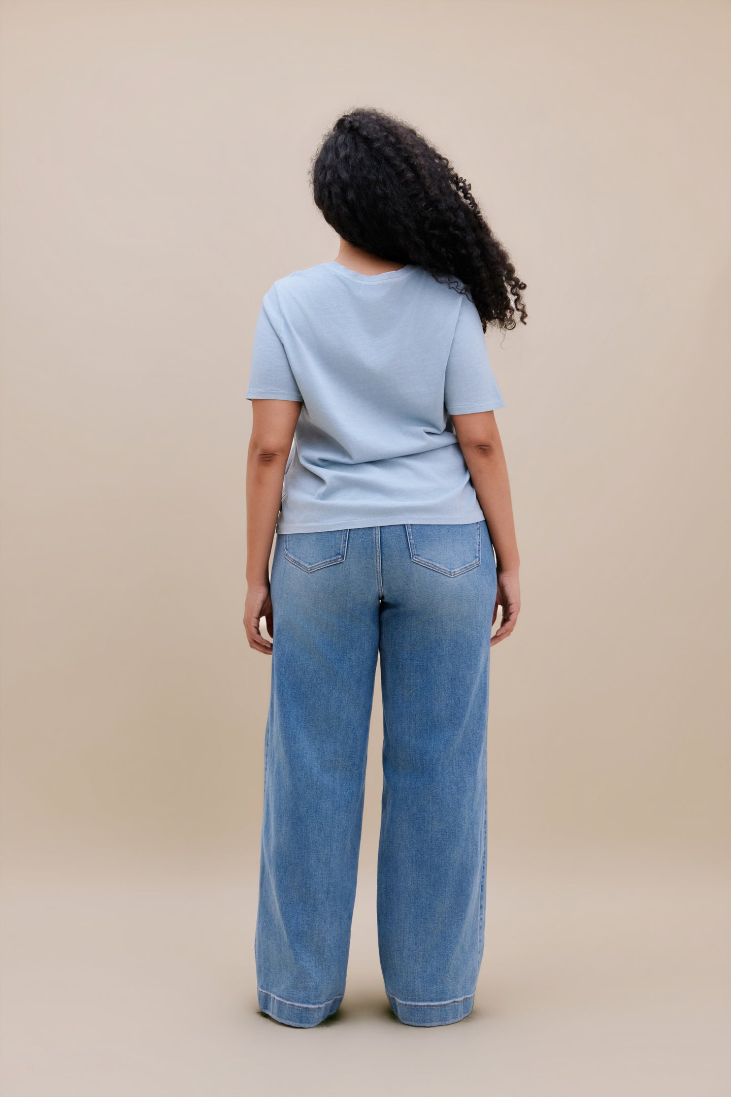Woman wearing a blue t-shirt and wide leg jeans with a beige background
