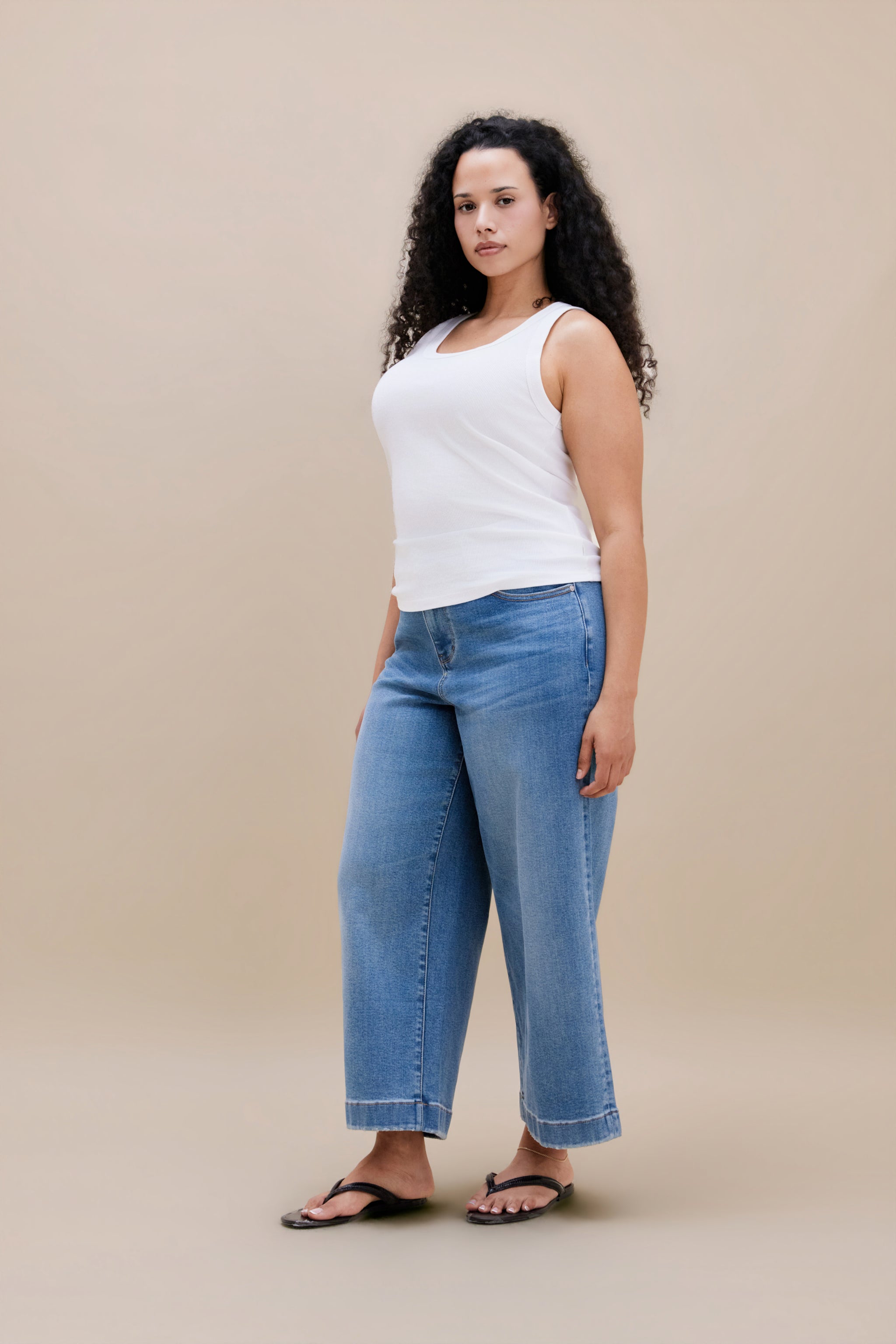 Woman wearing a white tank top with wide leg cropped jeans with a beige background 