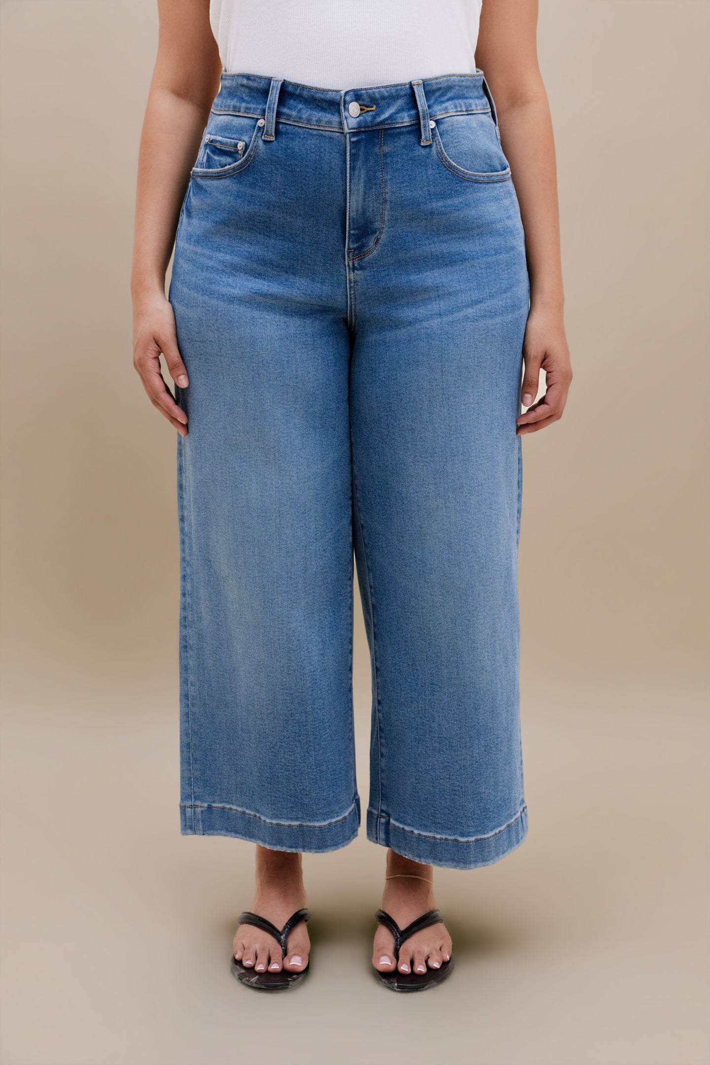 Bottom half of a woman wearing a white tank top with wide leg cropped jeans with a beige background 