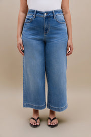 Bottom half of a woman wearing a white tank top with wide leg cropped jeans with a beige background 
