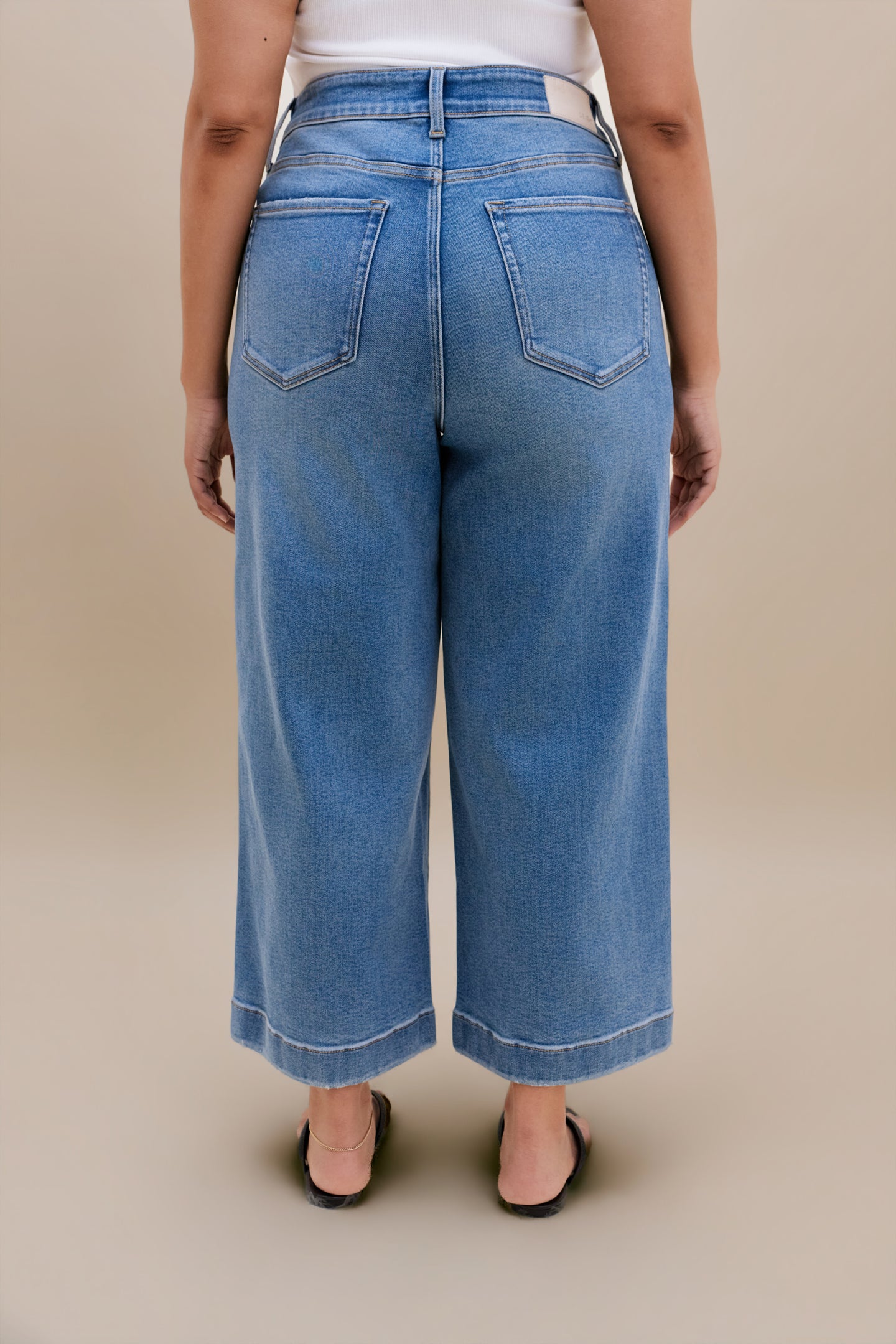Backside bottom half of a woman wearing a white tank top with wide leg cropped jeans with a beige background 