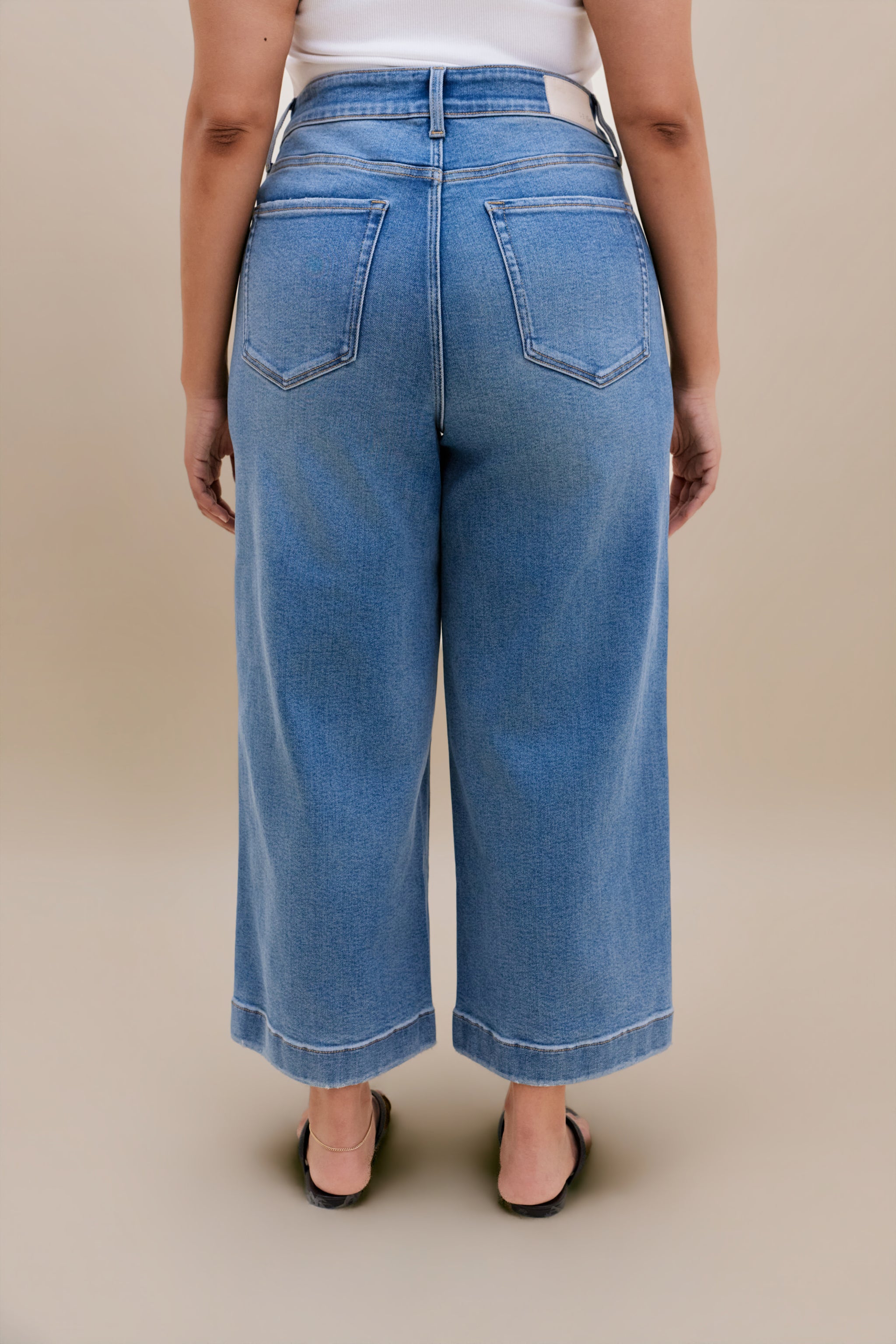 Backside bottom half of a woman wearing a white tank top with wide leg cropped jeans with a beige background 