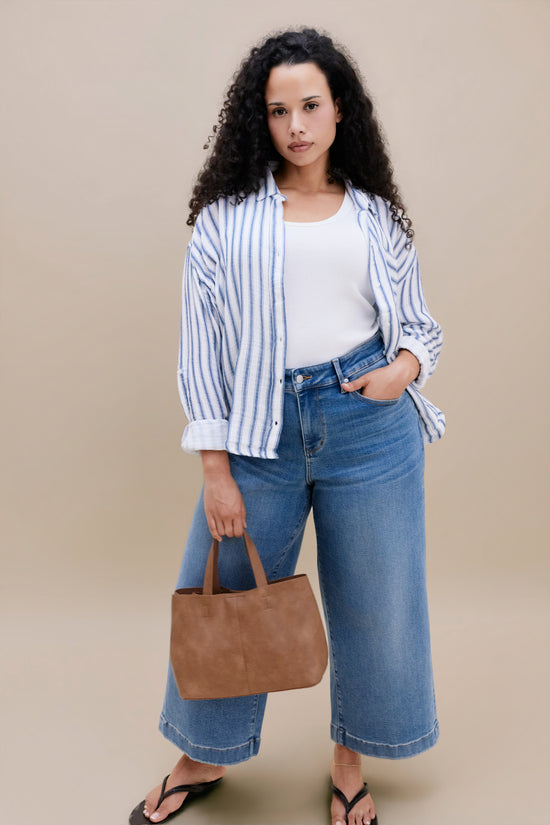 Woman wearing a white tank top and stripped blue button down shirt with wide leg cropped jeans with a beige background 