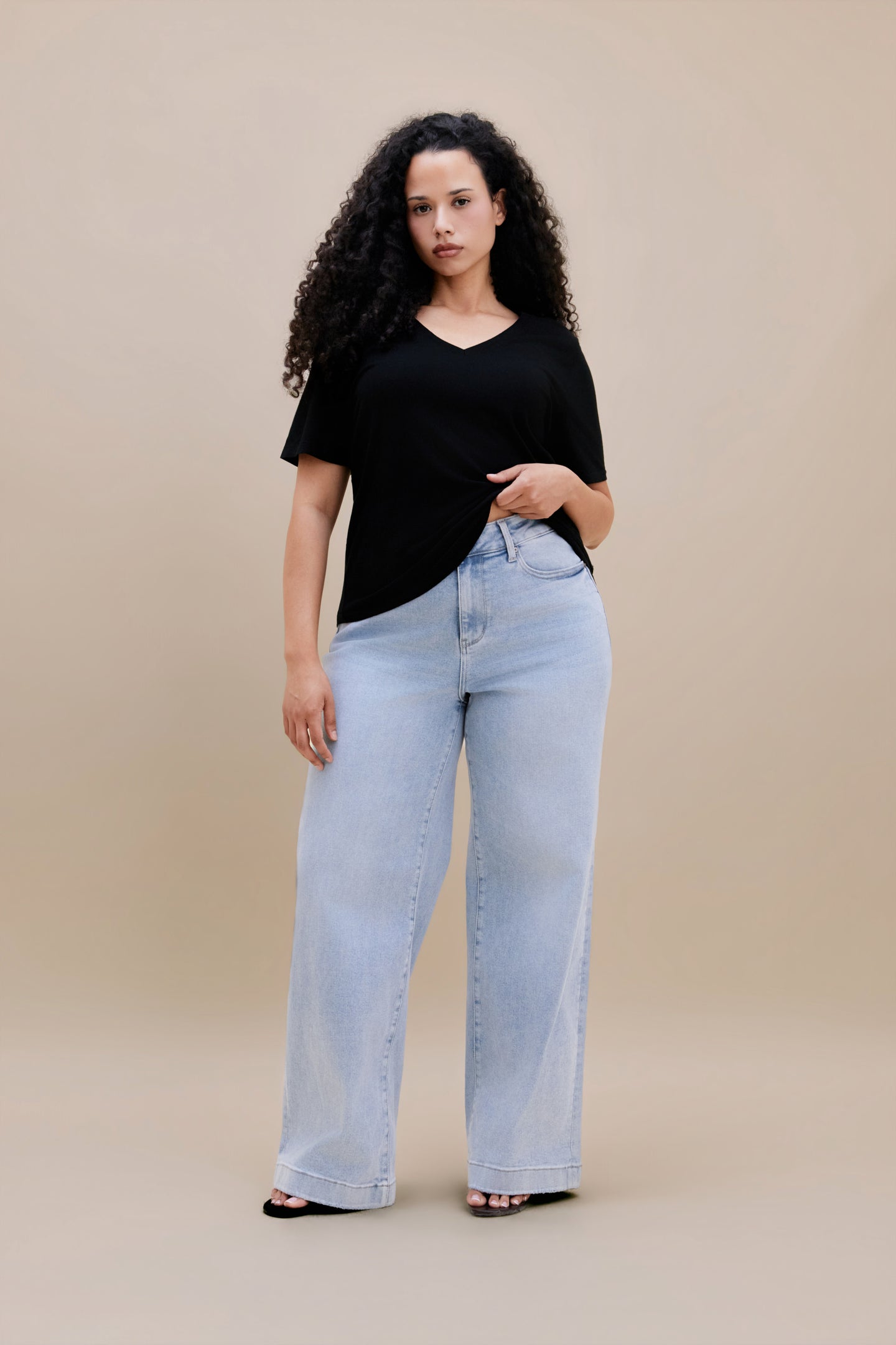 Woman wearing a black v-neck t-shirt and light colored wide leg jeans with a beige background | model-size: 16