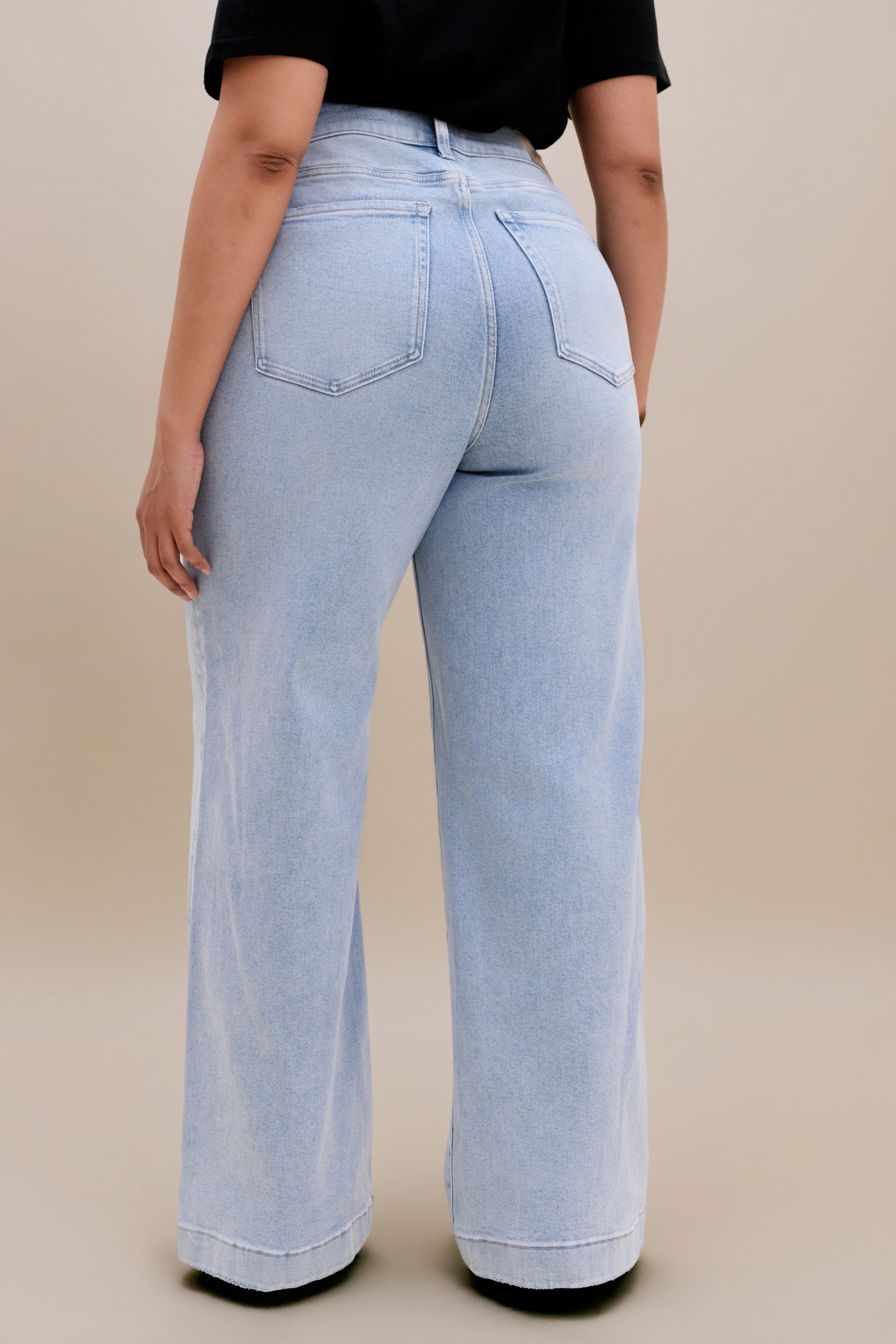 Backside bottom half of a woman wearing a black v-neck t-shirt and light colored wide leg jeans with a beige background 