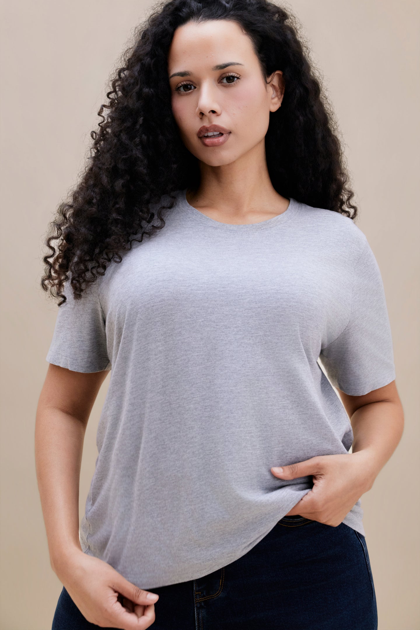 Woman wearing a light heather grey t-shirt  & dark jeans against a beige background