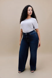 Woman wearing a gray t-shirt and dark wide leg jeans standing with a beige background
