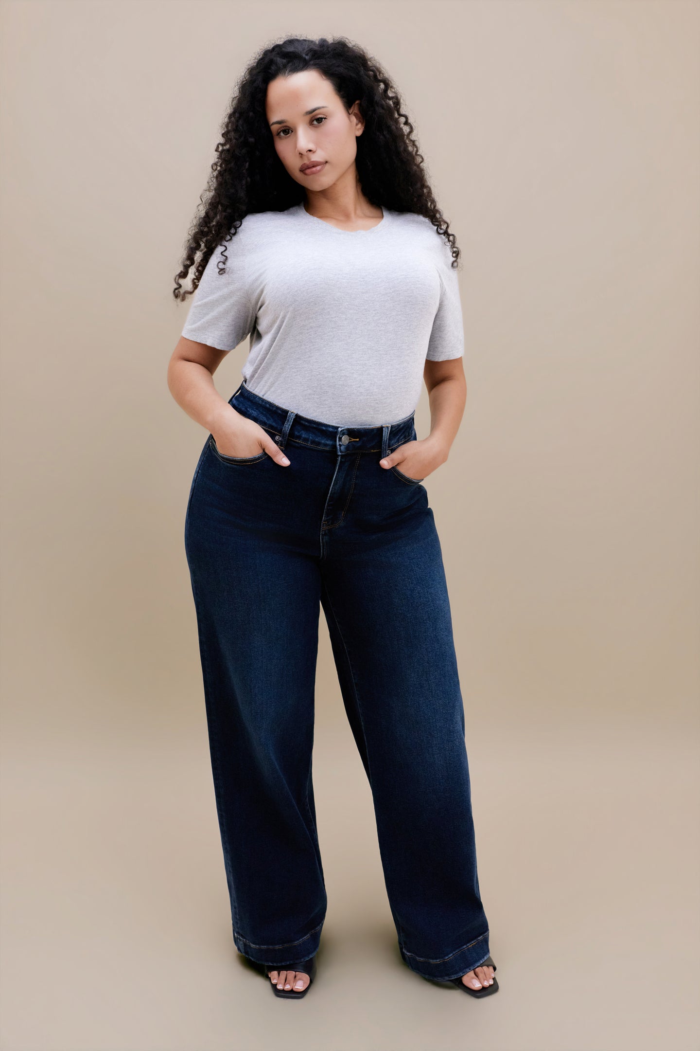 Woman wearing a light heather grey t-shirt  & dark jeans against a beige background