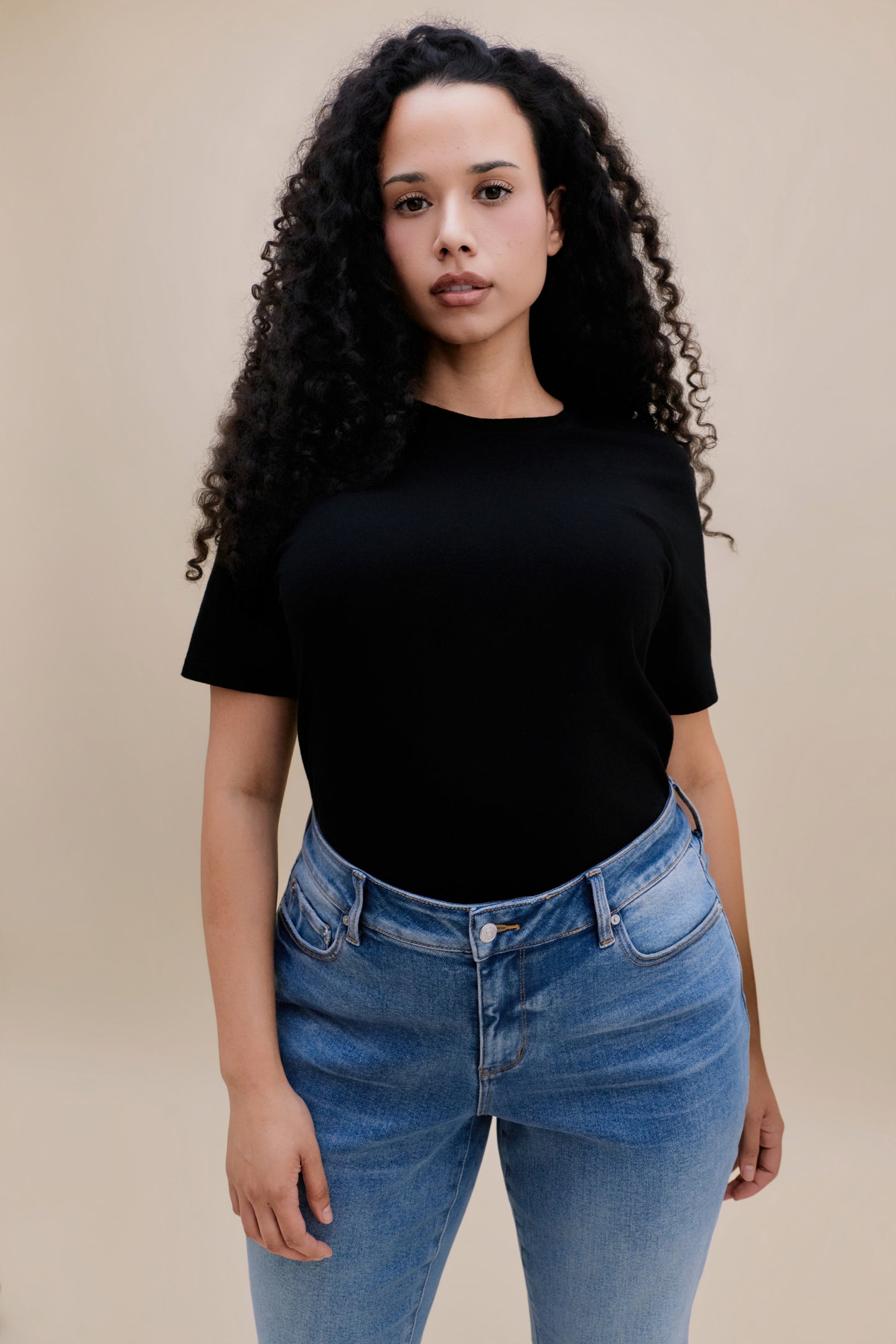 Woman wearing a black t-shirt and blue jeans against a beige background 