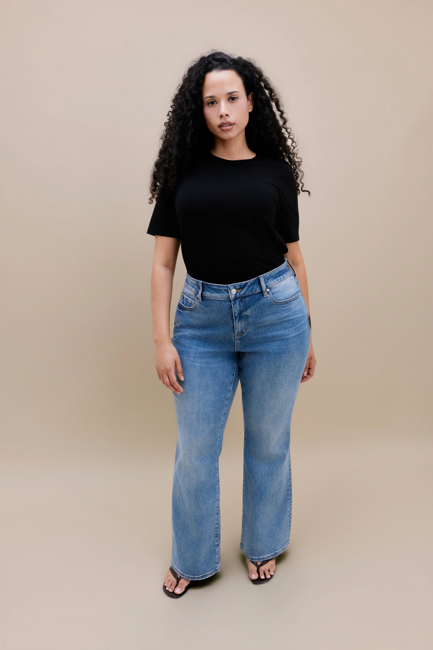 Woman wearing black t-shirt and bootcut jeans with a beige background | model size: 16