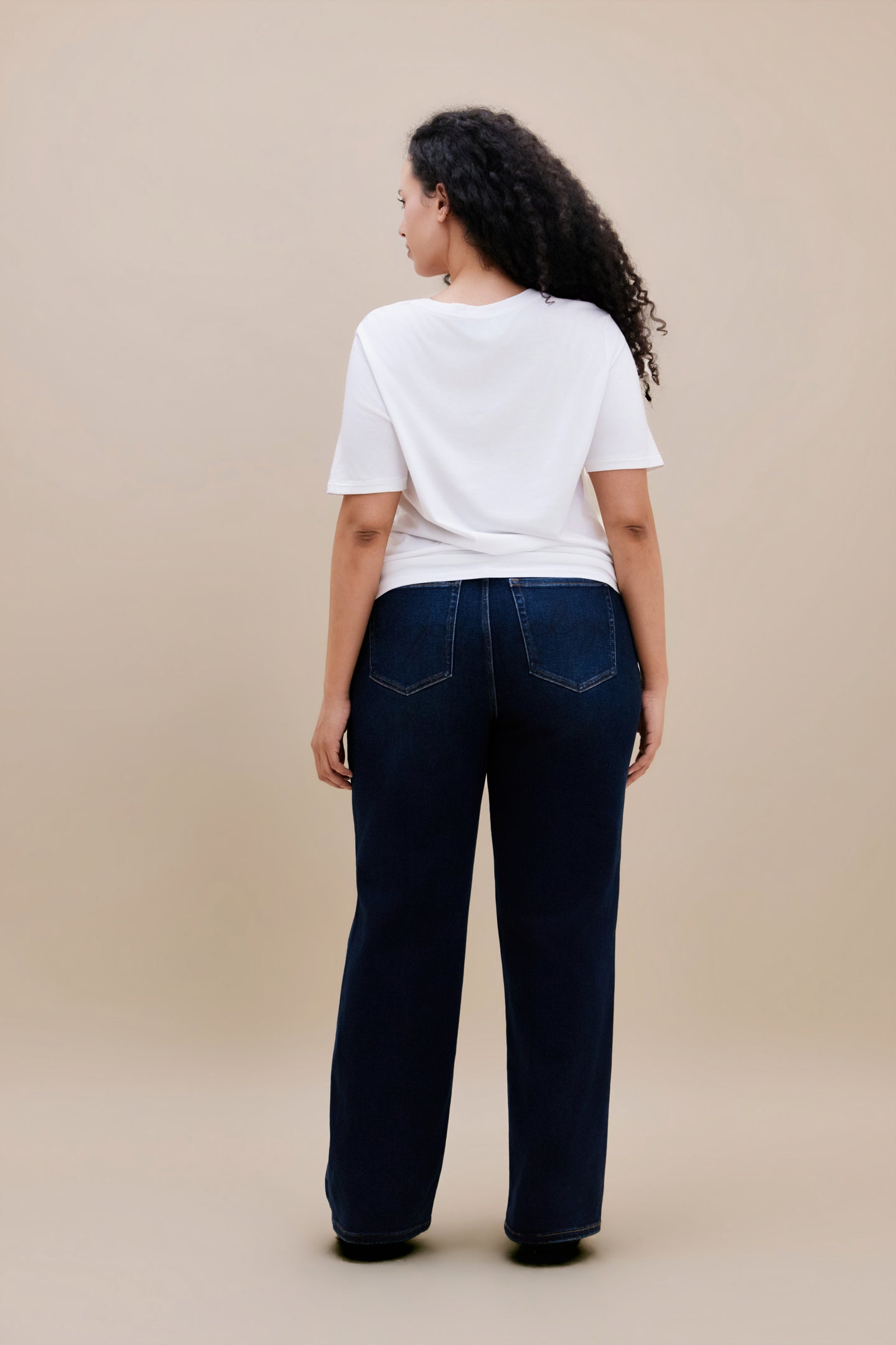 Woman wearing white t-shirt and dark straight leg jeans with a beige background