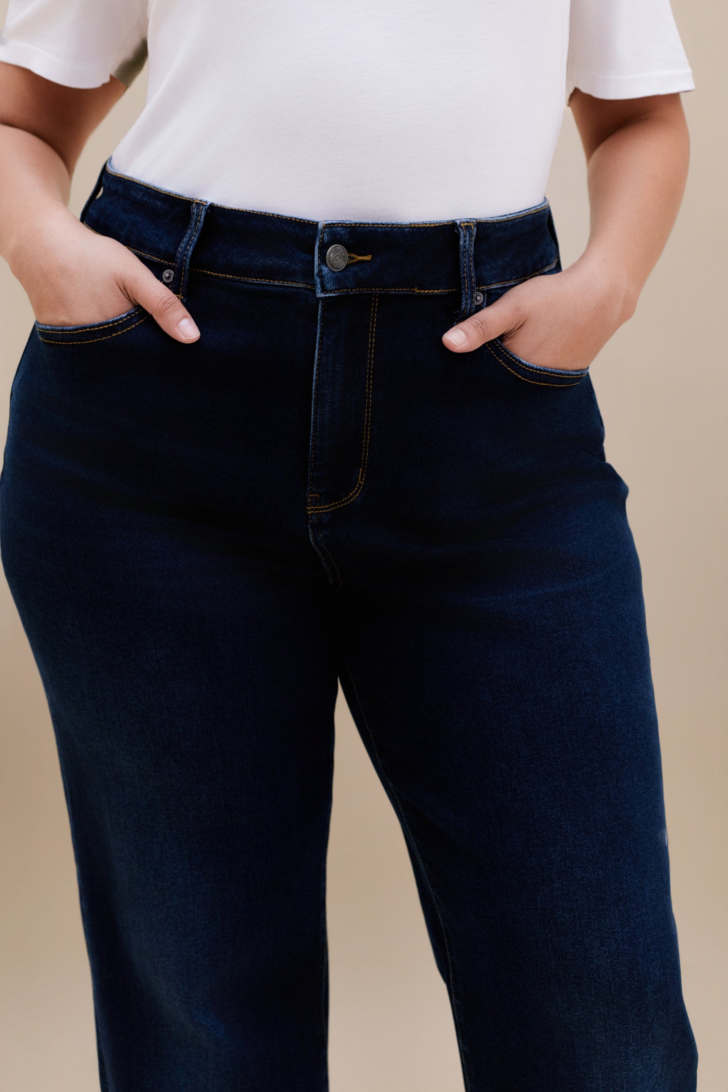 Close up highlighting the waist of a woman wearing white t-shirt and dark straight leg jeans with a beige background