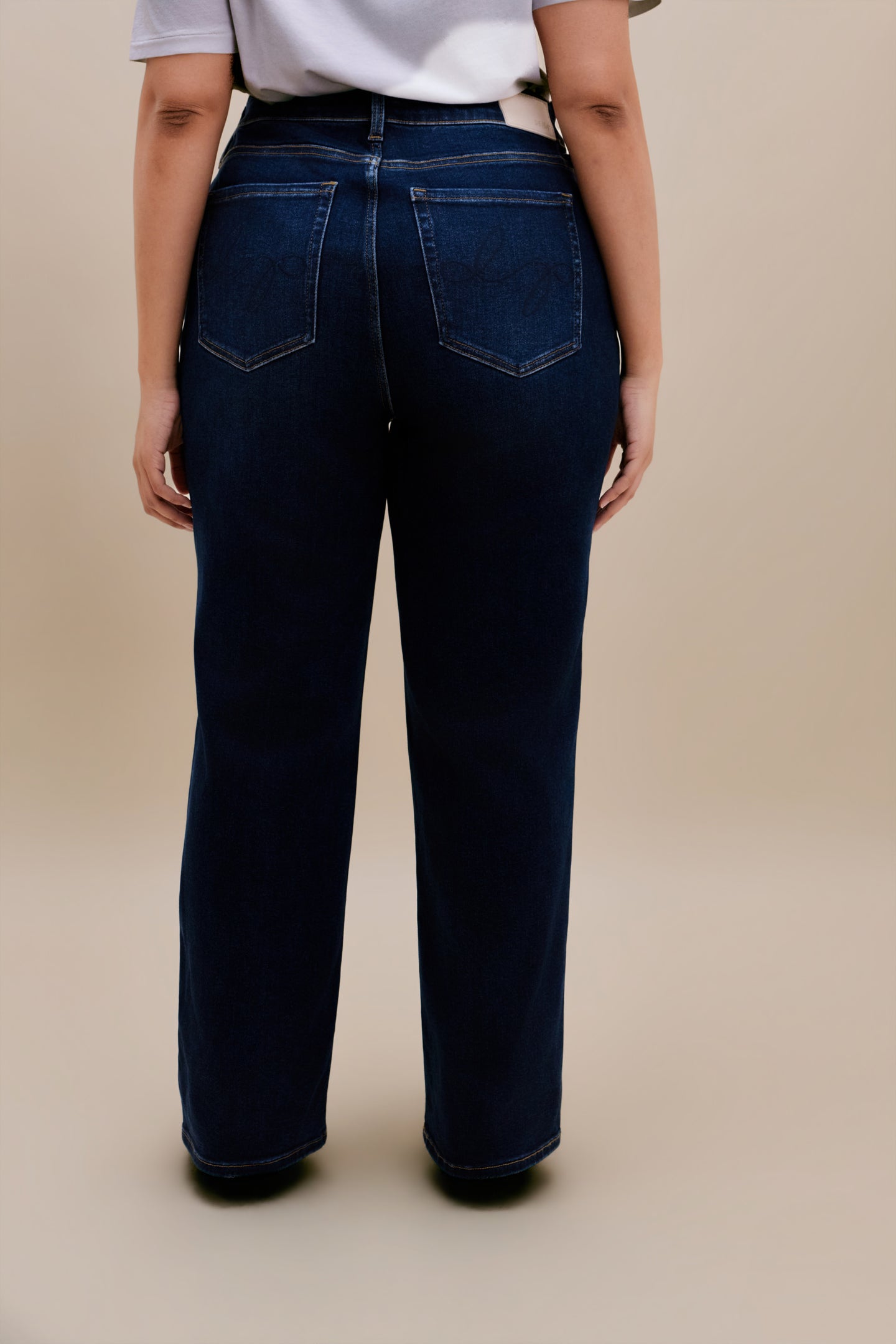Backside of a woman wearing white t-shirt and dark straight leg jeans with a beige background