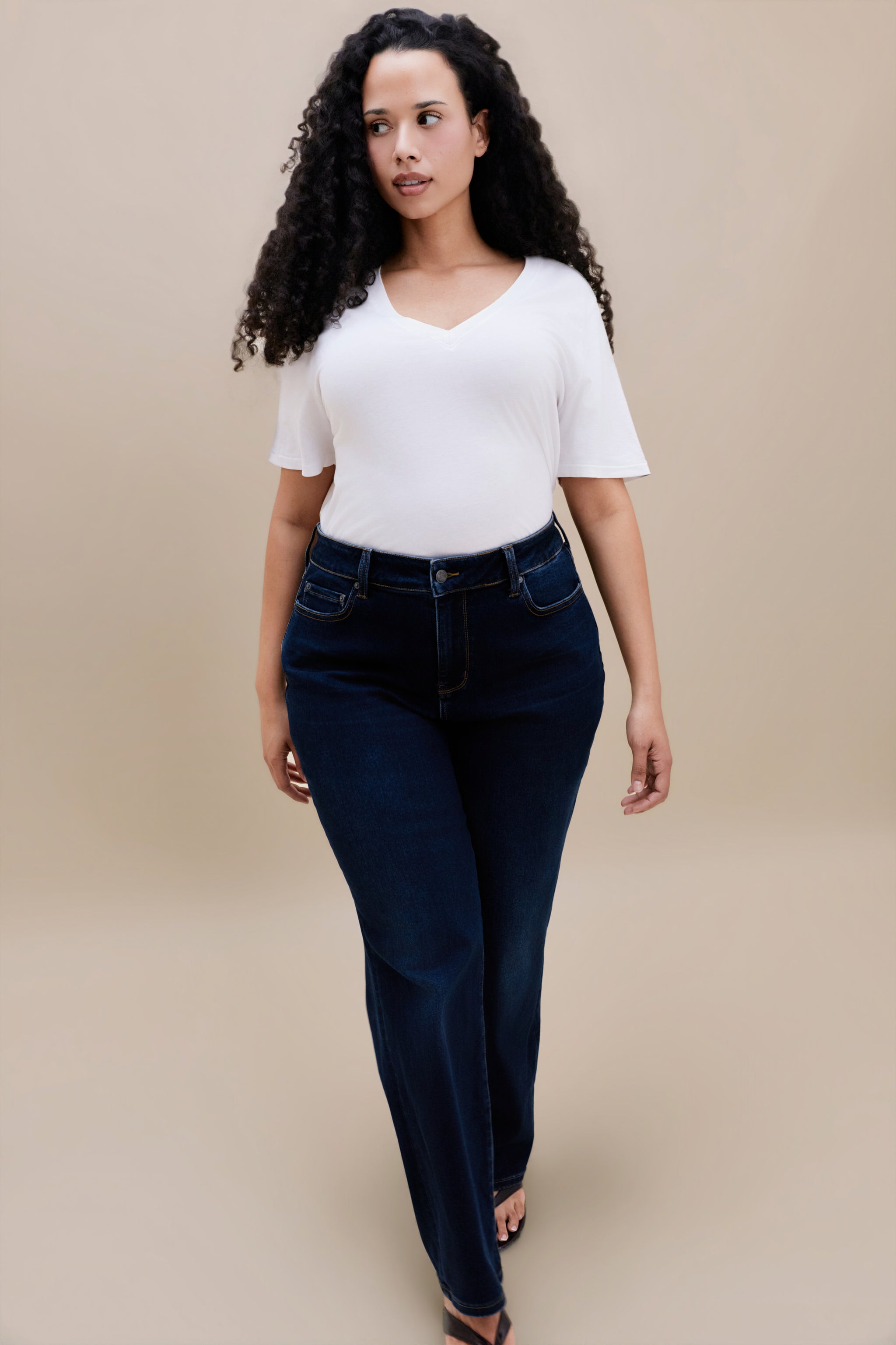 Woman wearing white t-shirt and dark straight leg jeans with a beige background