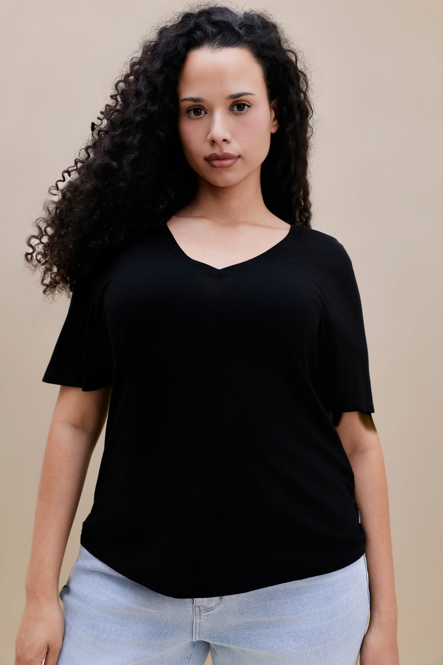 Close up of a woman wearing a black v-neck t-shirt and light blue jeans against a beige background