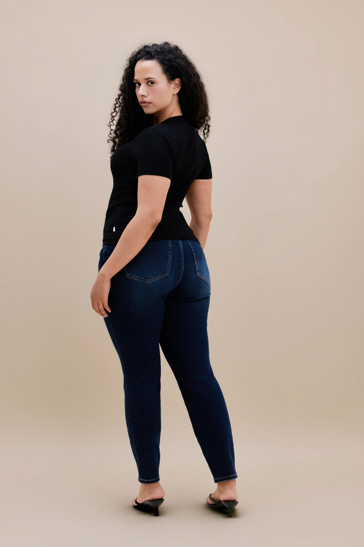 Backside of a woman wearing a black t-shirt and dark skinny jeans with a beige background 