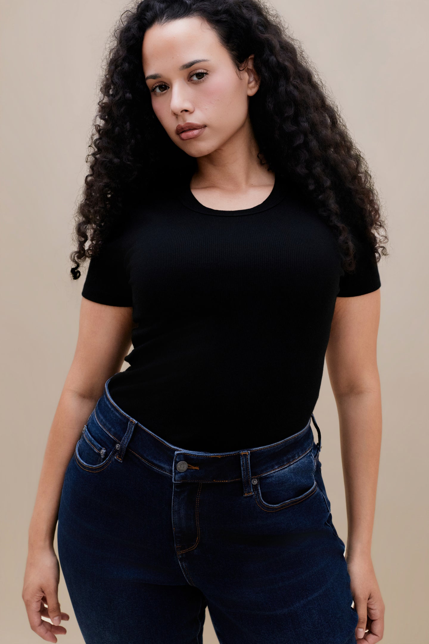 Close up of a woman wearing a fitted black rib tee and dark jeans against a beige wall 