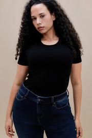 Close up of a woman wearing a fitted black rib tee and dark jeans against a beige wall 