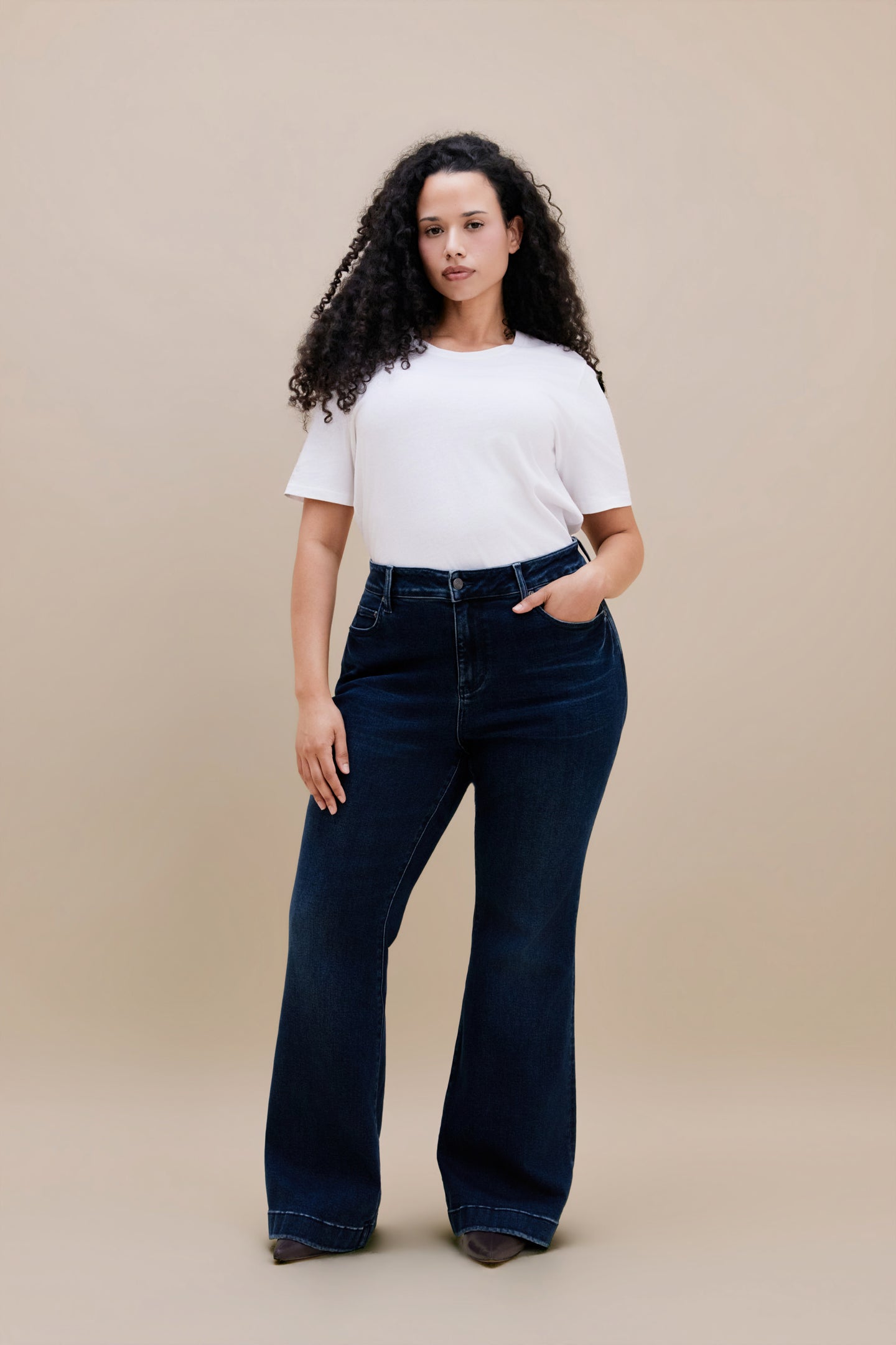 Woman wearing a white t-shirt and dark flare jeans with a beige background