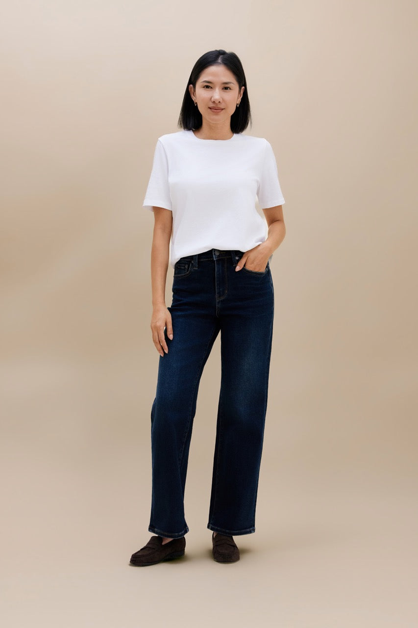A woman modeling a white short-sleeve crewneck t-shirt and dark blue wide-leg jeans, standing against a solid beige studio background.