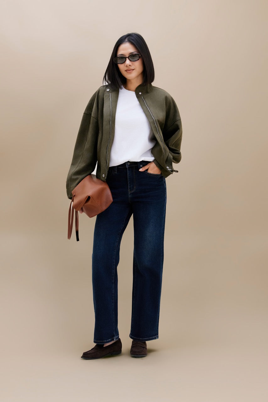 A woman modeling an olive green bomber jacket over a white t-shirt, paired with dark blue jeans, brown loafers, and a brown leather shoulder bag, set against a solid beige studio background.