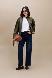 A woman modeling an olive green bomber jacket over a white t-shirt, paired with dark blue jeans, brown loafers, and a brown leather shoulder bag, set against a solid beige studio background.