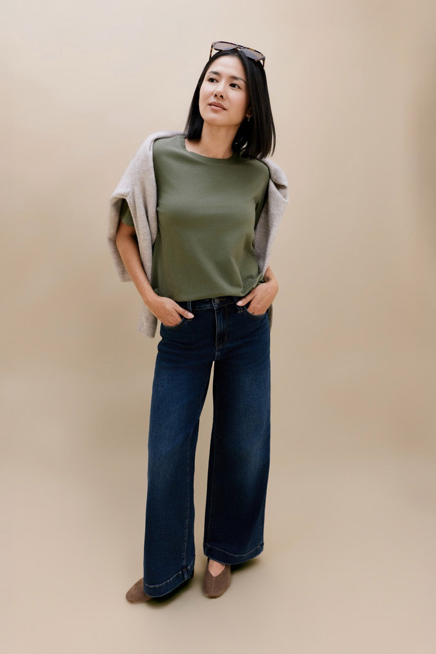 A woman modeling a sage green crewneck top with a light beige cardigan draped off her shoulders, paired with dark blue wide-leg jeans and brown flats, against a solid beige studio background.
