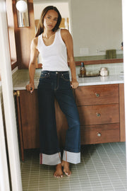 A woman stands barefoot in a bathroom doorway wearing dark blue wide-leg cuffed jeans with a white ribbed tank top. The bathroom has wood cabinets, a tiled floor, and a mirror behind her.