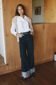 A woman stands indoors against a wood-paneled wall wearing a white button-up shirt, dark wide-leg jeans with rolled cuffs, a light beige cardigan draped over her shoulders, and black flats.