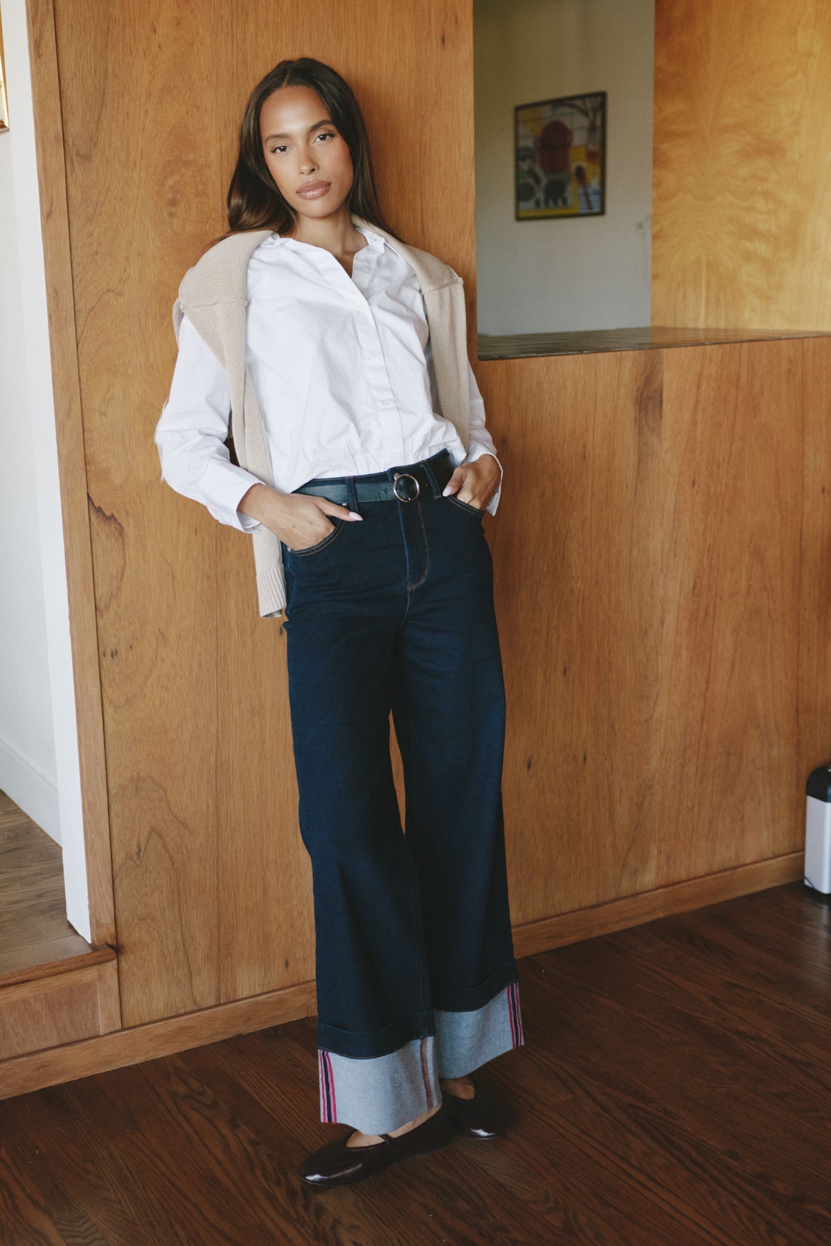 A woman stands indoors against a wood-paneled wall wearing a white button-up shirt, dark wide-leg jeans with rolled cuffs, a light beige cardigan draped over her shoulders, and black flats.
