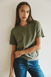 A woman wearing a muted olive green short-sleeve crewneck t-shirt and blue jeans, posing against a plain off-white wall.