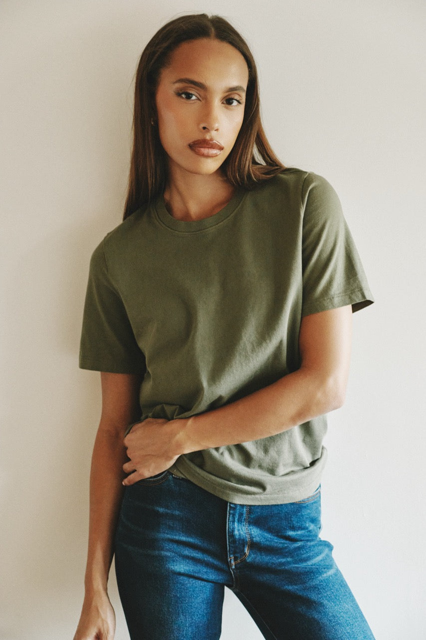 A woman wearing a muted olive green short-sleeve crewneck t-shirt and blue jeans, posing against a plain off-white wall.