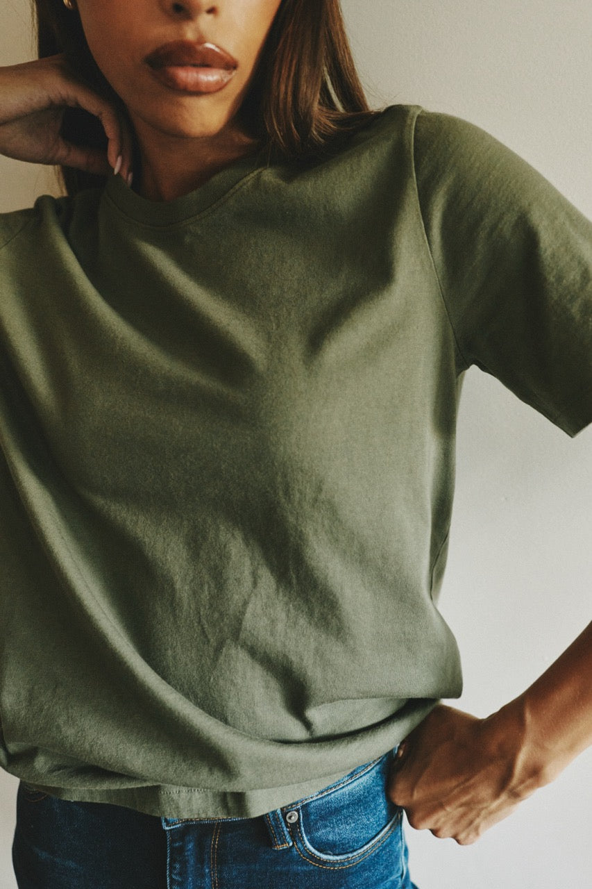 A close-up of a person wearing an olive green short-sleeve crewneck t-shirt tucked into blue jeans, with one hand on the hip and the other resting near the neck.