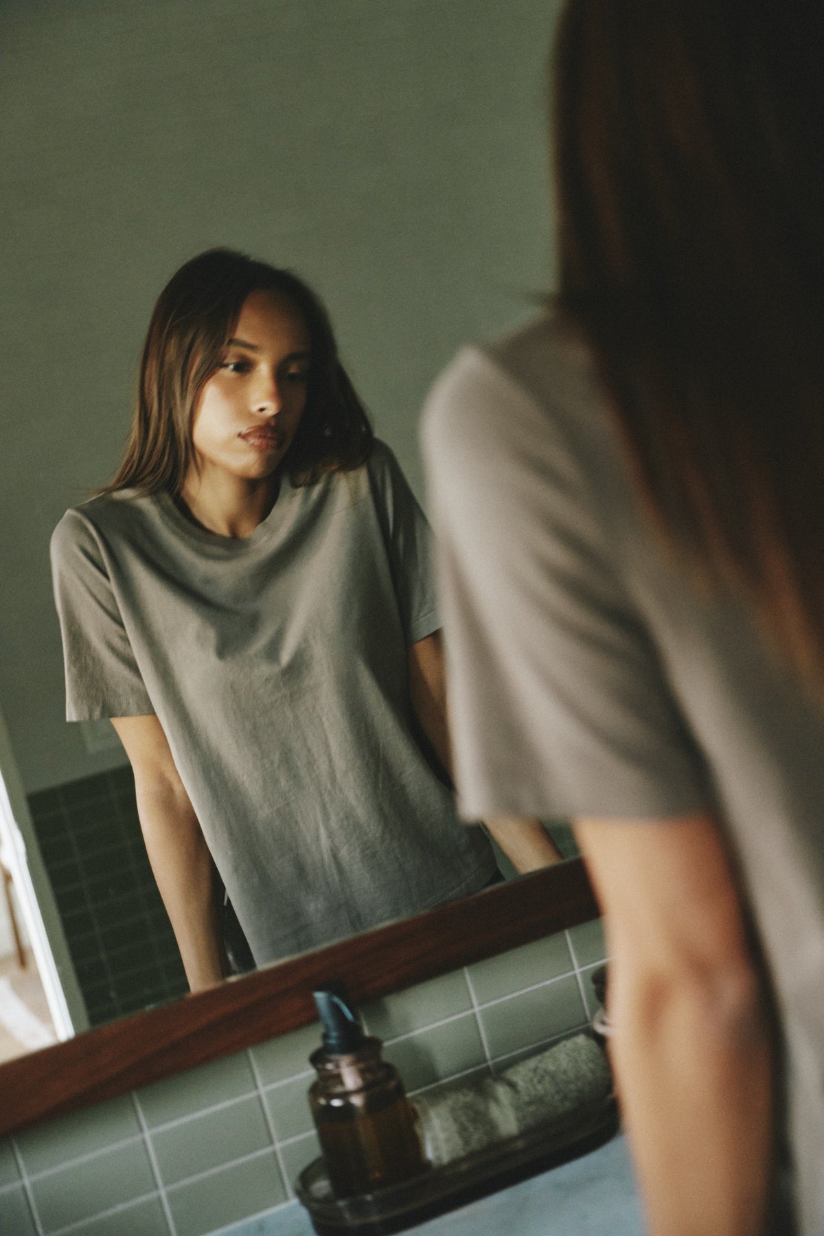 A woman wearing a muted gray t-shirt stands in front of a bathroom mirror, looking at her reflection. The bathroom has a tiled wall and a wooden-framed mirror.