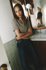 A woman with long dark hair stands in a tiled bathroom wearing a muted olive-gray short-sleeve t-shirt and dark blue jeans, with a sink and mirror visible behind her.
