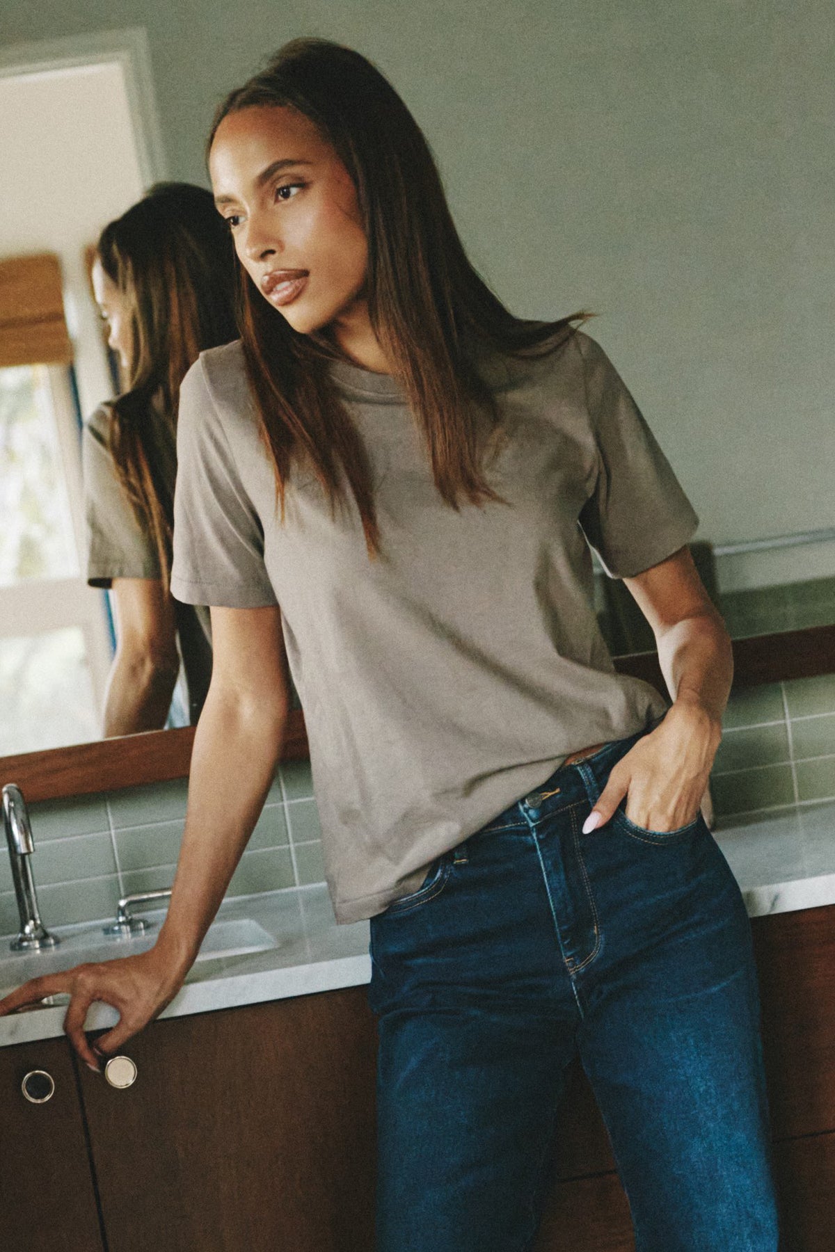 Woman wearing a taupe t-shirt and dark jeans leaning against a bathroom sink and staring out the window.