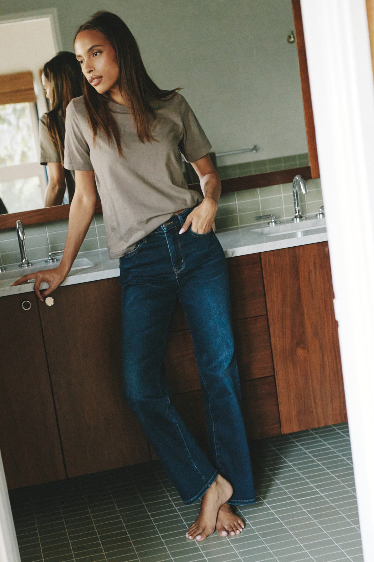 A woman stands barefoot in a bathroom wearing a taupe short-sleeve t-shirt and dark blue high-waisted flare jeans.